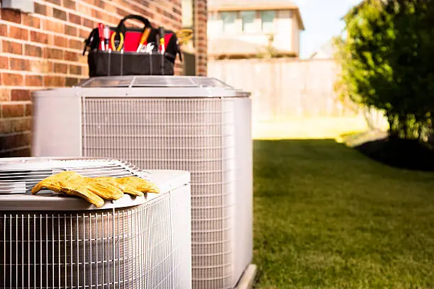 Two residential air conditioning condensers on a pad beside a brick wall, tool bag and work gloves resting on the units